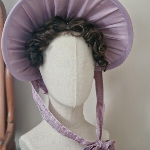 May include: A lavender bonnet with a wide, pleated brim and matching ribbons. The bonnet is displayed on a mannequin head with dark brown curly hair. The ribbons are tied in a bow under the chin.