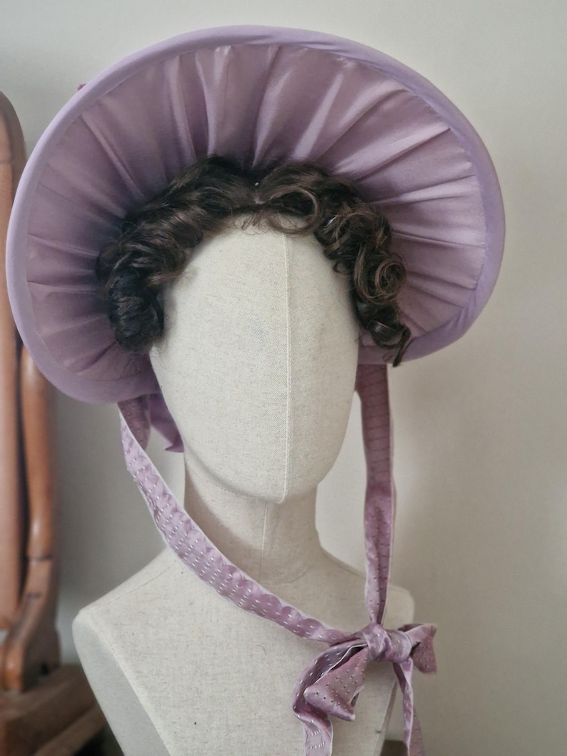 May include: A lavender bonnet with a wide, pleated brim and matching ribbons. The bonnet is displayed on a mannequin head with dark brown curly hair. The ribbons are tied in a bow under the chin.