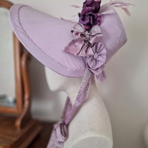 May include: A lavender-colored bonnet with a wide brim, adorned with dark purple fabric flowers, light purple feathers, and a large bow. A matching ribbon is tied around the neck. The hat is displayed on a mannequin head.