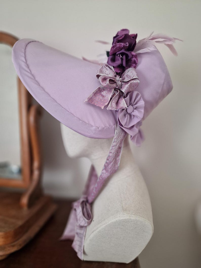 May include: A lavender-colored bonnet with a wide brim, adorned with dark purple fabric flowers, light purple feathers, and a large bow. A matching ribbon is tied around the neck. The hat is displayed on a mannequin head.