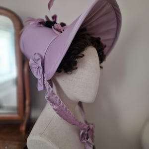 May include: A light purple bonnet with a wide brim, adorned with floral accents and a ribbon tie. The hat is displayed on a mannequin head, showcasing its design and shape. The bonnet's color is a soft, pastel hue, and the overall style is vintage.
