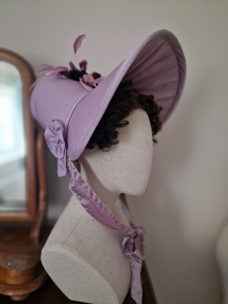May include: A light purple bonnet with a wide brim, adorned with floral accents and a ribbon tie. The hat is displayed on a mannequin head, showcasing its design and shape. The bonnet's color is a soft, pastel hue, and the overall style is vintage.