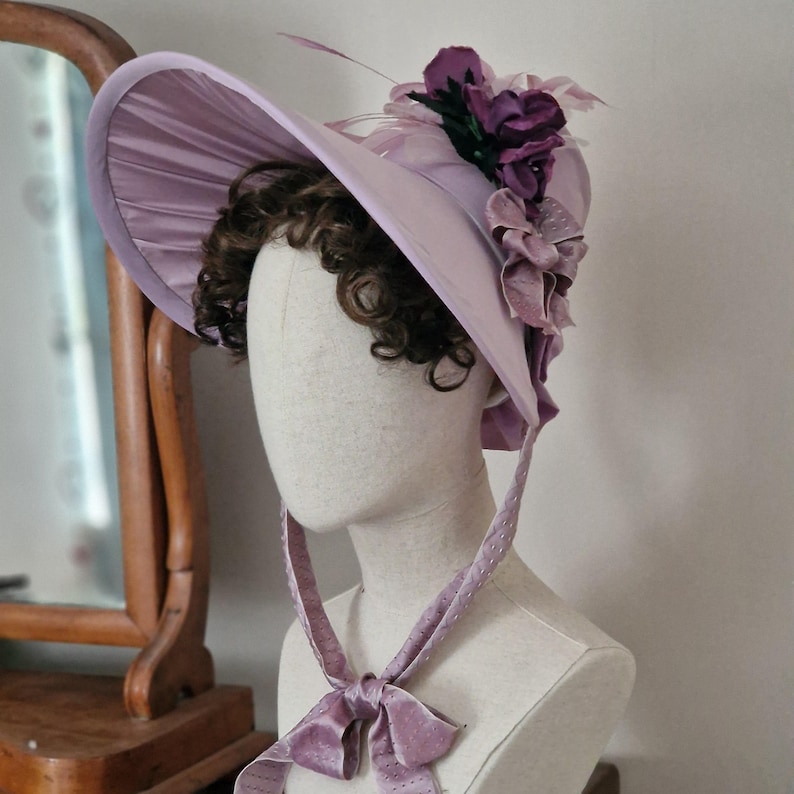 May include: A light purple bonnet with a wide brim and a decorative arrangement of purple flowers and feathers. The bonnet has a satin finish and is tied with a matching ribbon. The hat is displayed on a mannequin head.