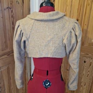May include: A beige cropped bolero jacket featuring long sleeves with gathered puff shoulders and a wide fold-over collar. The garment is constructed from a textured, woven fabric and is displayed on a red adjustable dress form mannequin.
