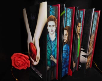 May include: A collection of books from the Twilight series, featuring cover art with character portraits and a red rose. The books are arranged with their spines visible, showcasing the titles and author's name.