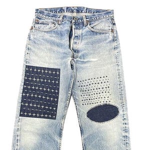 Può includere: Jeans di denim blu chiaro con toppe blu scuro e dettagli di cuciture a punto croce bianche. I jeans hanno un classico design a cinque tasche e una chiusura con bottoni.