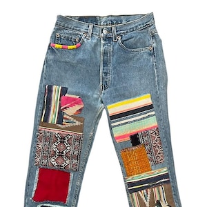 Op de afbeelding: Lichtblauwe spijkerbroek met kleurrijke stoffen patches op de voorkant genaaid. Patches bevatten verschillende patronen en texturen, zoals strepen, geometrische ontwerpen en haakwerk. De tailleband heeft een regenboogafwerking.