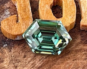 May include: A green emerald cut gemstone with a hexagonal shape, set on a wooden surface with the word "JOM" spelled out in wooden letters.