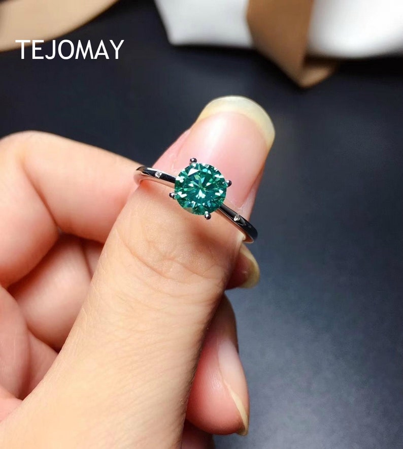 May include: A silver ring with a green gemstone set in a prong setting. The ring is on a finger.