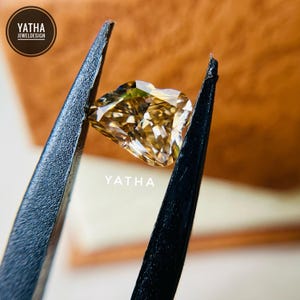 May include: A heart-shaped, champagne-colored diamond is held by black tweezers. The diamond has multiple facets that reflect light. The word "YATHA" is printed below the diamond. The image also includes a logo that says "YATHA JEWELDESIGN".
