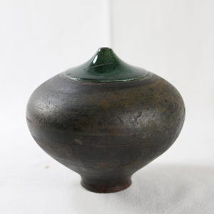 May include: A handcrafted ceramic vase with a dark, textured body and a glossy green top. The vase has a rounded shape with a narrow neck and a dark brown base. This unique piece is perfect for home decor.
