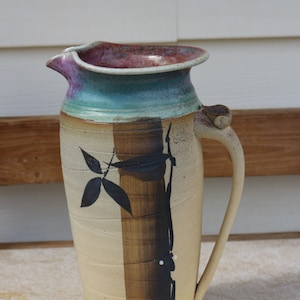 May include: A ceramic pitcher with a brown, black, and white design. The pitcher has a wide mouth and a handle. The handle is attached to the side of the pitcher.