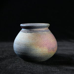 May include: A small, handcrafted ceramic pot with a rounded body and a narrow neck. The pot features a textured surface with a blend of colors, including blue, purple, and gold. Two dark lines encircle the neck. The pot sits on a black surface.