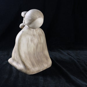 May include: A ceramic figurine of a person sitting with their head down, wearing a long robe. The figurine is a light brown color and has a smooth, polished finish.