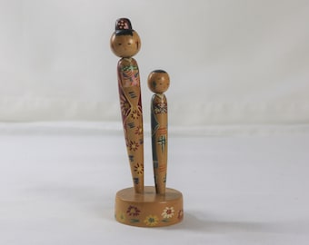 May include: Two wooden dolls, one taller than the other, stand on a base with painted flowers. The dolls are painted with colourful clothing and faces.