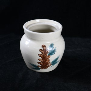 May include: A white ceramic jar with a lid, featuring a hand-painted design of a brown pine cone with green pine needles.