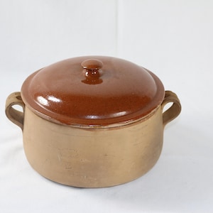 May include: A brown ceramic cooking pot with a matching lid. The pot has two handles and a rounded shape. The lid has a small knob on top. The pot is a light tan color, and the lid is a glossy brown.