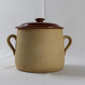 May include: A tan ceramic pot with a brown lid and two handles. The pot has text etched into the side. The lid has a small knob. The pot is likely used for cooking or storage.