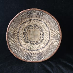 May include: A round, woven basket with a brown rim and intricate geometric patterns in shades of beige and dark brown. The basket's design features concentric circles and zigzag patterns, creating a textured surface.