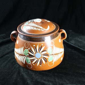May include: A brown ceramic pot with a lid, decorated with white and green floral designs. The pot has two handles and a brown rim.