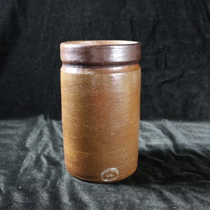 May include: A cylindrical, brown stoneware jar with a dark brown rim. The jar has a textured surface and a small, circular marking near the base. The background is a dark, textured fabric.