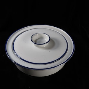May include: A round white ceramic casserole dish with a matching lid. The dish and lid feature a smooth white finish accented by thin, dark blue circular bands along the edges. A small, circular knob handle sits in the center of the lid.