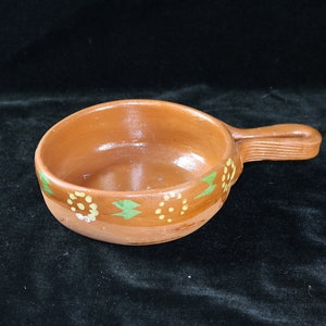 May include: A small, brown terracotta bowl with a handle. The bowl is decorated with green and yellow painted flowers.