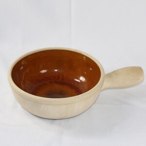 May include: A ceramic soup bowl with a handle. The interior is a glossy brown, contrasting with the off-white exterior. The bowl is round with a single handle, suitable for serving soups or stews.
