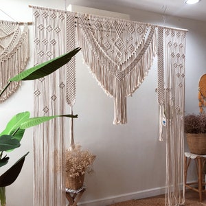May include: A set of three macrame wall hangings in a cream color. The hangings are made of cotton cord and feature geometric patterns. The hangings are displayed on a white wall with a plant in the foreground.