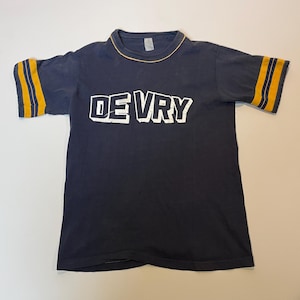 May include: Navy blue t-shirt with the word "DE VRY" in white block letters. The shirt has gold and navy blue striped sleeves and a gold trim around the collar. The shirt is made of a soft, worn material.