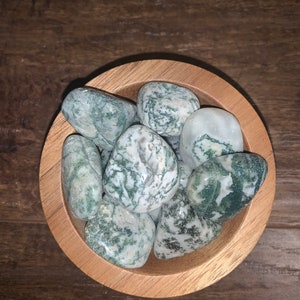 May include: A wooden bowl filled with moss agate tumbled stones. The stones are a mix of green and white with a swirling pattern.