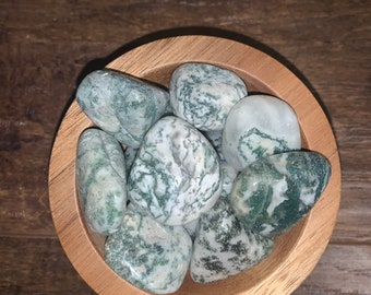May include: A wooden bowl filled with moss agate tumbled stones. The stones are a mix of green and white with a swirling pattern.