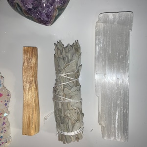 May include: A set of four items for smudging and cleansing rituals. The set includes a white selenite crystal, a bundle of white sage, a palo santo stick, and a cluster of amethyst crystals.
