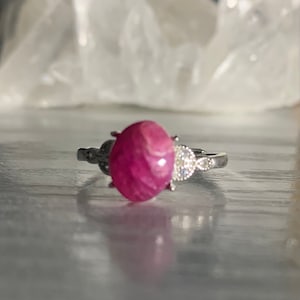 May include: A silver ring with a pink oval gemstone and smaller clear gemstones on the band.