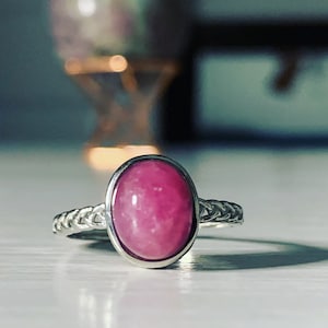 May include: A silver ring with a pink oval gemstone set in a silver bezel. The ring band is braided.