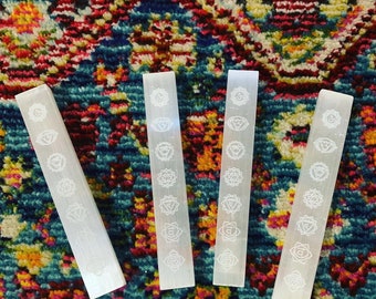 May include: Four white selenite wands with the seven chakra symbols etched on each. The wands are arranged vertically on a colorful rug.
