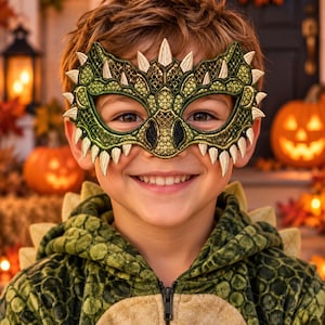May include: A green and gold embroidered dragon mask featuring a textured scale pattern, pointed spikes along the edges, and black accents around the eye openings. The mask is designed to cover the upper face and is worn with a matching green hoodie.