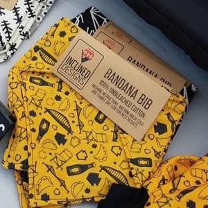 May include: A yellow bandana bib with a black and white print of various objects, including chairs, bottles, and a bridge. The bib is made of 100% unbleached cotton and has the text "BANDANA BIB" and "INCLINED DESIGNS" printed on it.