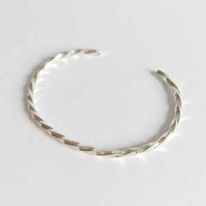 Könnte beinhalten: Ein silbernes, gedrehtes Armkettchen. Das Armband besteht aus einem einzigen Stück Metall, das zu einer Spirale gedreht ist.