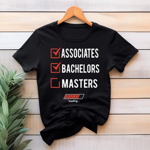 Peut inclure: T-shirt noir avec texte blanc et cases à cocher rouges. Le texte indique "ASSOCIATES", "BACHELORS" et "MASTERS", avec des coches à côté des deux premiers. Un graphique de barre de chargement est en bas.