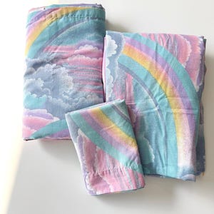 May include: A set of folded bed linens featuring a repeating pattern of pastel rainbows, fluffy pink and blue clouds, and soft yellow sunbeams. The fabric displays a whimsical sky-themed print with a light blue background and soft color palette.