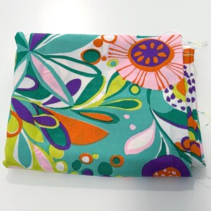 May include: A folded piece of fabric featuring a vibrant floral pattern on a teal background. The design includes large stylized flowers in shades of pink, purple, and orange, accented by green leaves and small circular shapes throughout the print.