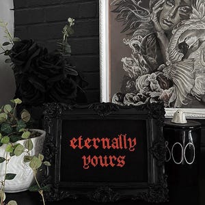 May include: A framed cross-stitch piece with the words "eternally yours" in red lettering. The frame is black and ornate. A black ceramic jar with "boo" written on it is to the right of the frame. Black roses and greenery are also in the image.