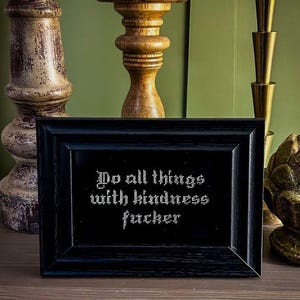 May include: A black framed picture with white text that reads "Do all things with kindness fucker" on a wooden surface with candlesticks.