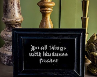 Pode incluir: Uma imagem emoldurada em preto com texto branco que diz "Do all things with kindness fucker" sobre uma superfície de madeira com castiçais.