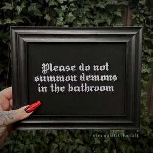 May include: A black framed cross-stitch with white text that reads "Please do not summon demons in the bathroom". The cross-stitch is on a black fabric background.