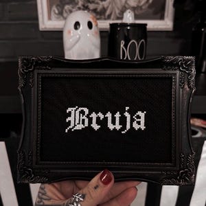 May include: Black framed cross-stitch art with the word "Bruja" in white gothic lettering. The frame has ornate detailing. A small white ghost figurine and a black "BOO" jar are in the background, suggesting a Halloween theme.