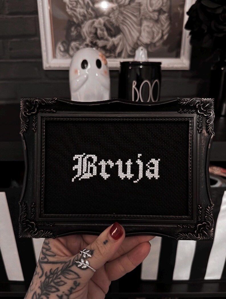 May include: Black framed cross-stitch art with the word "Bruja" in white gothic lettering. The frame has ornate detailing. A small white ghost figurine and a black "BOO" jar are in the background, suggesting a Halloween theme.