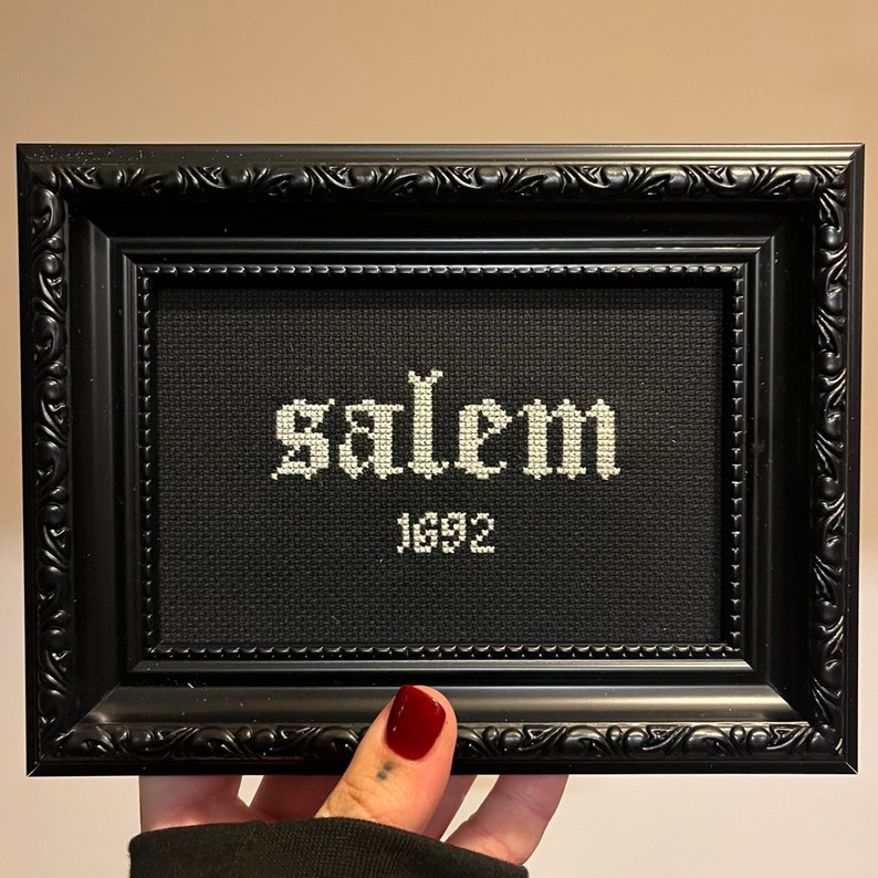 May include: A black framed cross-stitch with white embroidery that reads "Salem 1692".