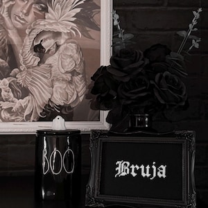 May include: A black and white gothic-themed display featuring a framed artwork, a black ceramic jar with "BOO" in white, a framed cross-stitch with the word "Bruja", and a bouquet of black roses. The background includes a black brick wall.